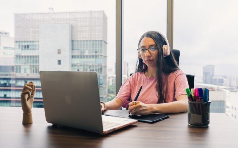 Woman working on laptop with headphones and stylus.