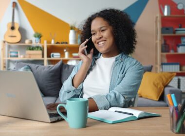 Woman talking on phone while working on laptop at desk.